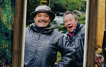 Bob and Paul stand in wet weather gear in front of a river