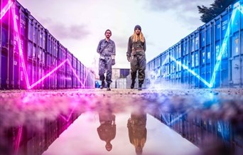 Two youths in boiler suits stand between shipping containers. Neon lighting zig-zags on the side of the containers.