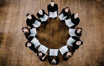 A choir, photographed from above, stand in a circle with their sheet music open.