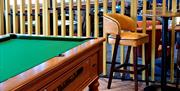 A pool table with herringbone style flooring, hanging plants, stools and a table to put drinks on