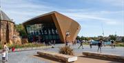 Firstsite Art Gallery in the sunshine, with a festival going on around it, people enjoying the sun, and playing basketball outside the front of the bu