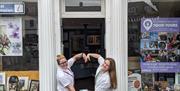 Two staff members form a heart with their arms in front of the Tourist Information Centre, to mark English Tourism Week