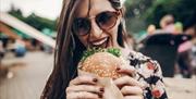 Women with sunglasses and long hair enthusiastically eating large burger