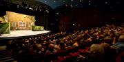 The inside of the auditorium at Queen's Theatre Hornchurch. A full auditorium is looking at a stage. The stage has a yellow house set with two hedges,