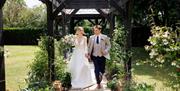 A bride and groom walking in gardens from a pergola