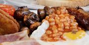 a cooked breakfast compromising of sausage, fried egg, baked beans, bacon, grilled tomatoes, mushrooms and hash brown