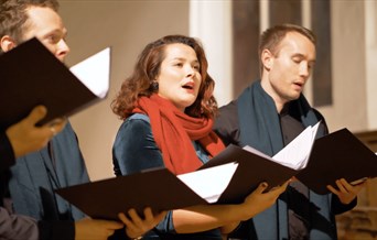 Two men and woman - wearing a red scarf - hold hymn books and sing as if in a choir.