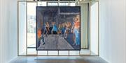 A large fresco painting by Rosie Hastings and Hannah Quinlan of women fighting hangs on a wooden frame covering the width of the room. Daylight comes
