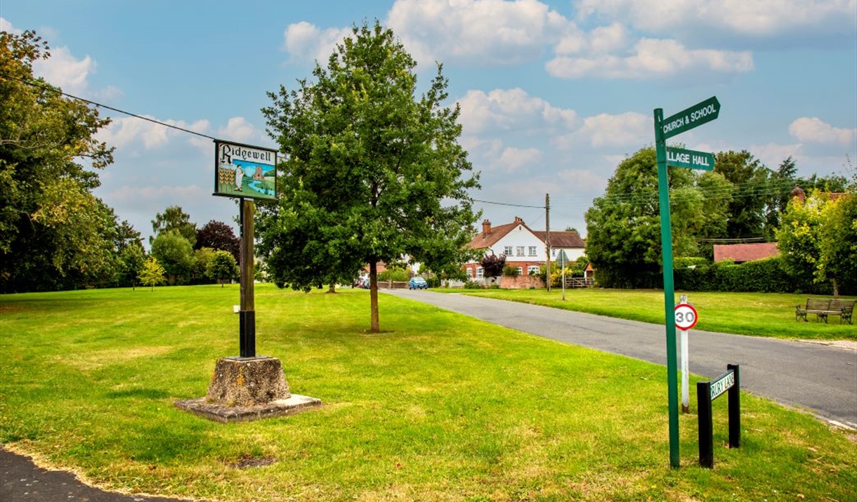 A village green, showing a Ridgewell village sign, a tree and a signpost to the School.