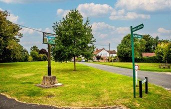 A village green, showing a Ridgewell village sign, a tree and a signpost to the School.