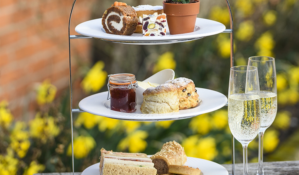 Close-up of a three-tier cake stand showing an afternoon tea; sandwiches/savoury pastries, scones with cream and jam, and a selection of cake.