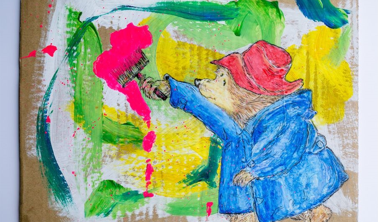 An artwork depicting Paddington Bear painting brightly coloured brush strokes on cardboard.