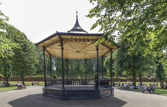 the victorian bandstand in castle park, surrounded by trees and people sitting on wooden benches. the bandstand has dark blue decorative ironwork and