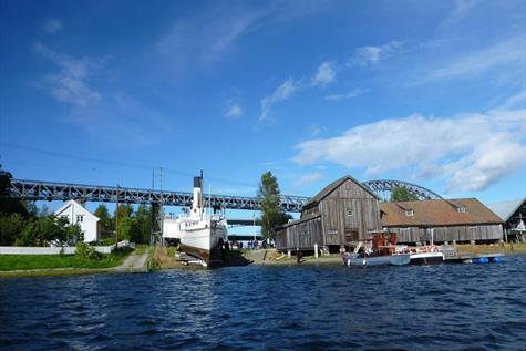 båter ved sjøen en sommerdag med broen og hvite hus samt naust i bakgrunnen. Mjøssamlingene på Minnesund
