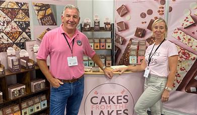 Owners of Cakes From The Lakes pose in pink t-shirts at a stand with cakes and treats