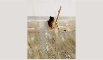 Women on the beach setting up an easel dressed in all white.