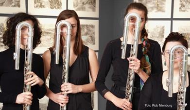 Four women dressed in black stand holding large U-shaped flutes (contrabass flutes) in front of their faces, partially obscuring them. They are standi
