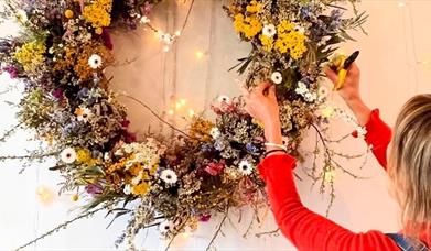 A person wearing a red long-sleeve shirt adjusts a large floral wreath made of colorful dried flowers, including yellow, white, and purple blooms, han