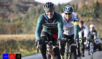 Group of cyclists riding on a country road, with two riders in green jerseys leading the way and smiling.