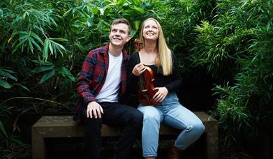 a young man and woman sitting on a wooden bench surrounded by dense green foliage