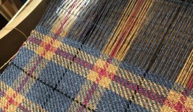 Close-up of a woven tartan fabric on a loom, featuring a pattern of blue, yellow, and red threads in a traditional plaid design