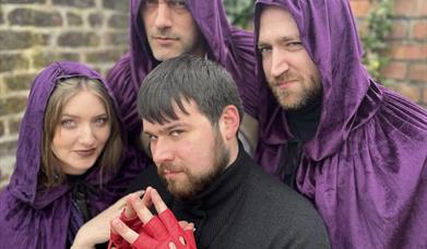 There are four people posing together. Three of them are wearing deep purple hooded cloaks the fourth is in black wearing red gloves