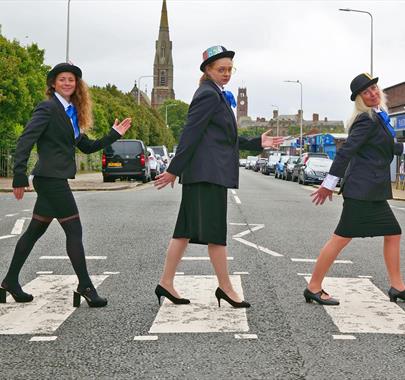 Three women dressed in black suits, blue ties, and bowler hats walking across a zebra crossing in a humorous recreation of the Beatles' Abbey Road pho