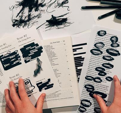 Hands holding printed pages with blackout poetry created using thick black marker, surrounded by other poem drafts, pens, tape, and scribbled paper on
