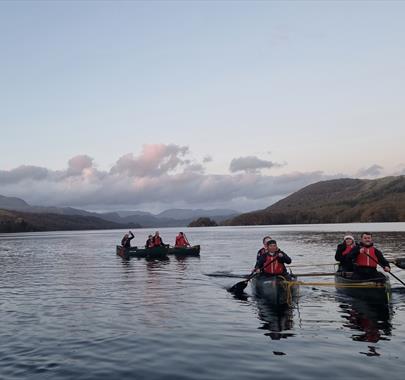 8 canoeists paddling on a calm lake at dusk in Cumbria, silhouetted against a soft, fading sky with hills in the distance