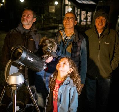 Two children looking through a telescope at night accompanied by 4 adults