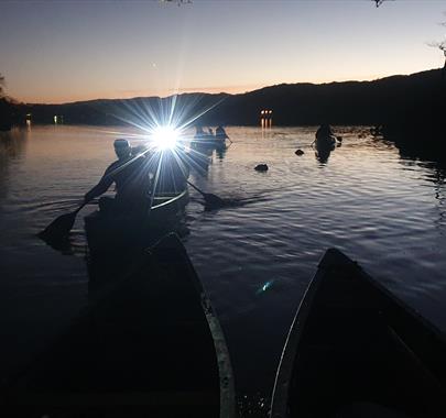 Canoeists on a lake in Cumbria at night, with a bright light from their canoe shining toward the camera, reflecting on the dark water