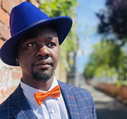Man wearing a bright blue hat, orange bow tie, and checkered suit standing beside a brick wall in a sunlit outdoor setting, looking thoughtfully into