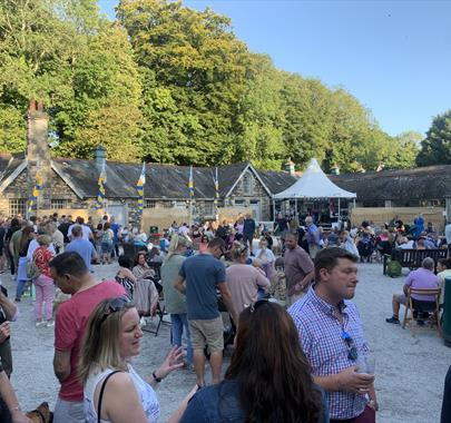 Large crowd gathered at an outdoor event with livemusic, food, and drinks. Set against a backdrop of historic stone buildings and trees on a sunny day
