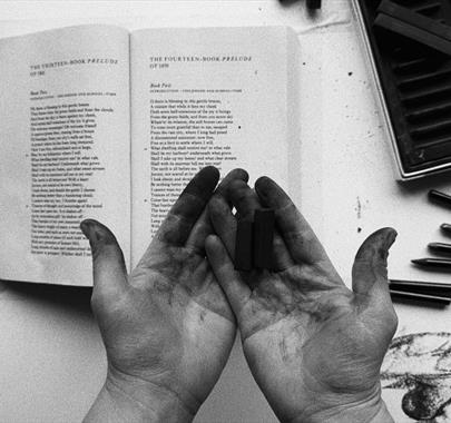 Black and white photo of dirty palms hovering above a table with a book and pencils