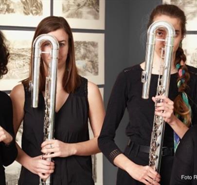 Four women dressed in black stand holding large U-shaped flutes (contrabass flutes) in front of their faces, partially obscuring them. They are standi