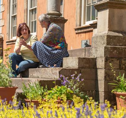 Amanda Thackeray sitting on the stone steps in a Georgian-style garden, speaking to another woman, surrounded by lush greenery.