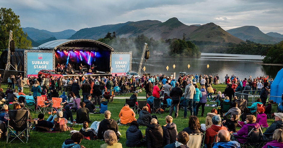 The Subaru stage at the Lake District festival is at the back. Behind there is a beautiful lake. People are sat down or standing waiting listening to the band on stage. 