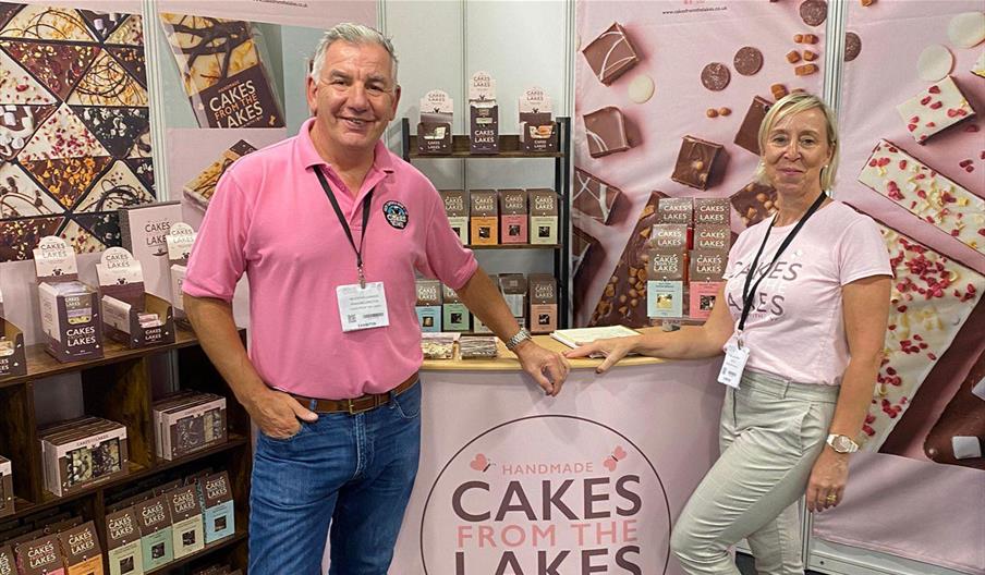 Owners of Cakes From The Lakes pose in pink t-shirts at a stand with cakes and treats