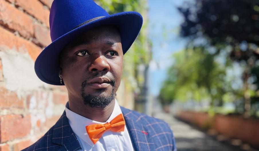 Man wearing a bright blue hat, orange bow tie, and checkered suit standing beside a brick wall in a sunlit outdoor setting, looking thoughtfully into