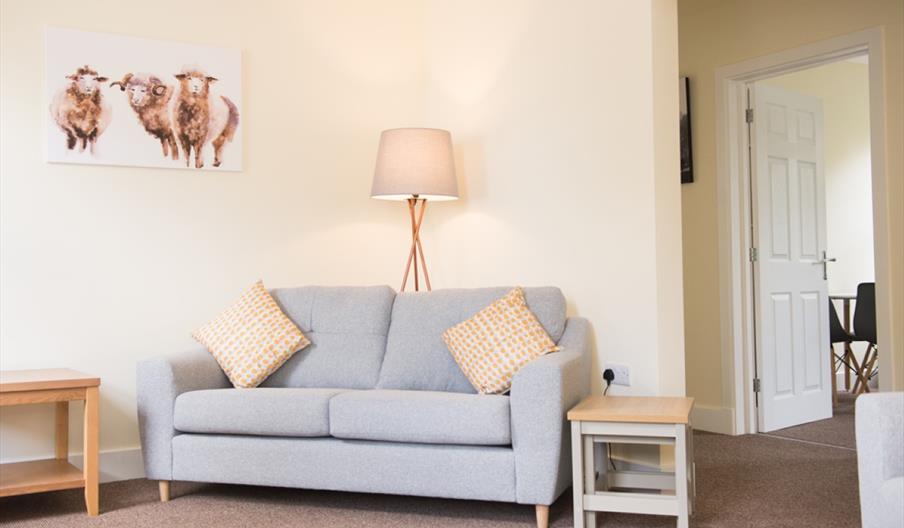 The lounge in one of the Park Hill Mews cottages showing a sofa, side tables, lighting and a picture of sheep on the wall.