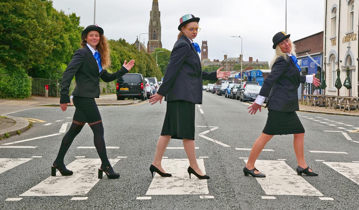 Three women dressed in black suits, blue ties, and bowler hats walking across a zebra crossing in a humorous recreation of the Beatles' Abbey Road pho