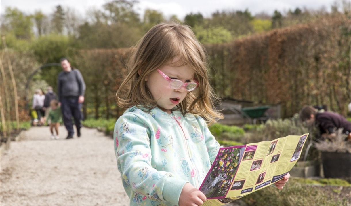 A young child stands on a gravel garden path holding an open, colourful leaflet. Behind them, several adults and children walk along the path, and a p