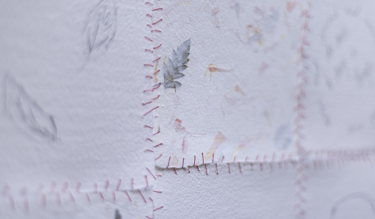 Close-up of layered white paper pieces stitched together with visible red thread, forming a patchwork surface with faint leaf prints and soft, texture
