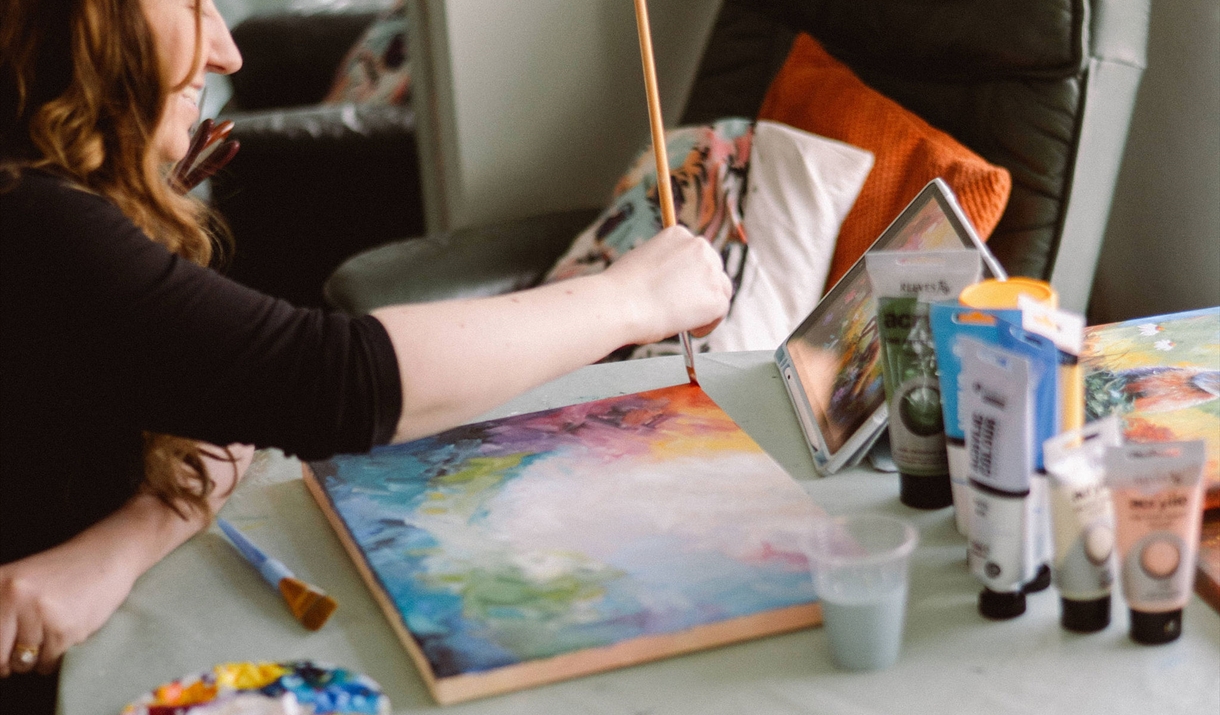 A person painting a colourful landscape on canvas with acrylic paints and a tablet beside them