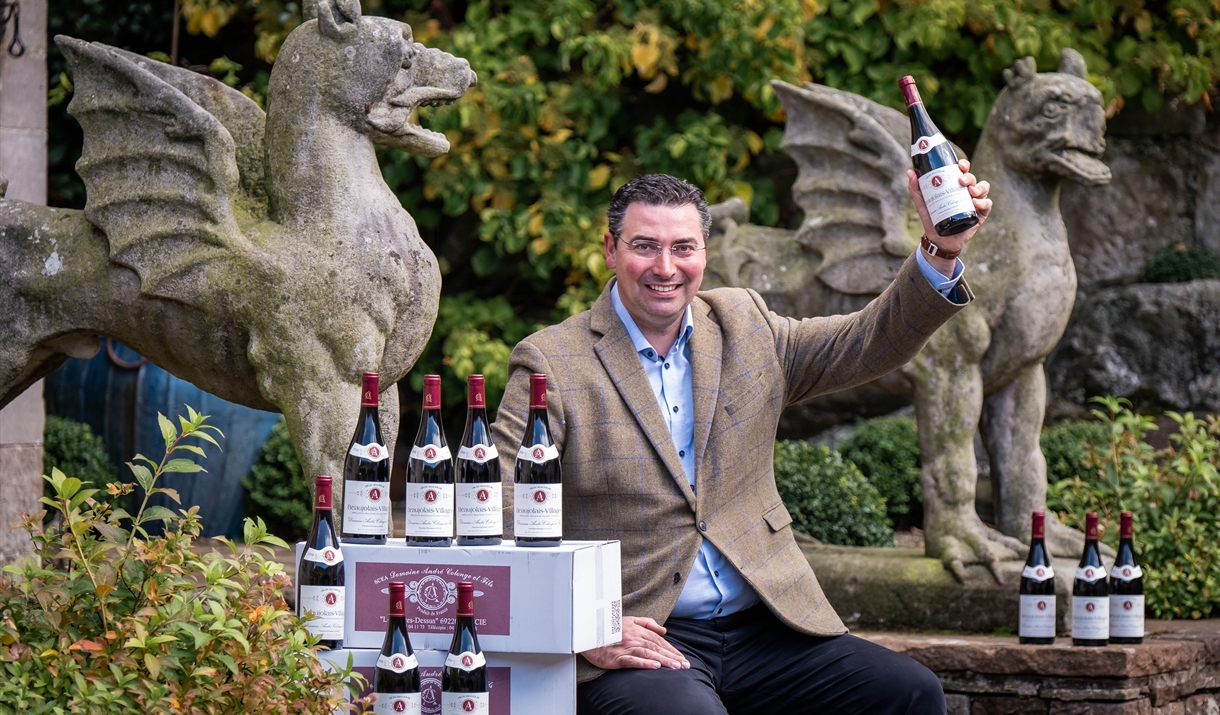 A smiling man holding up a bottle of red wine sat next to two stone statues