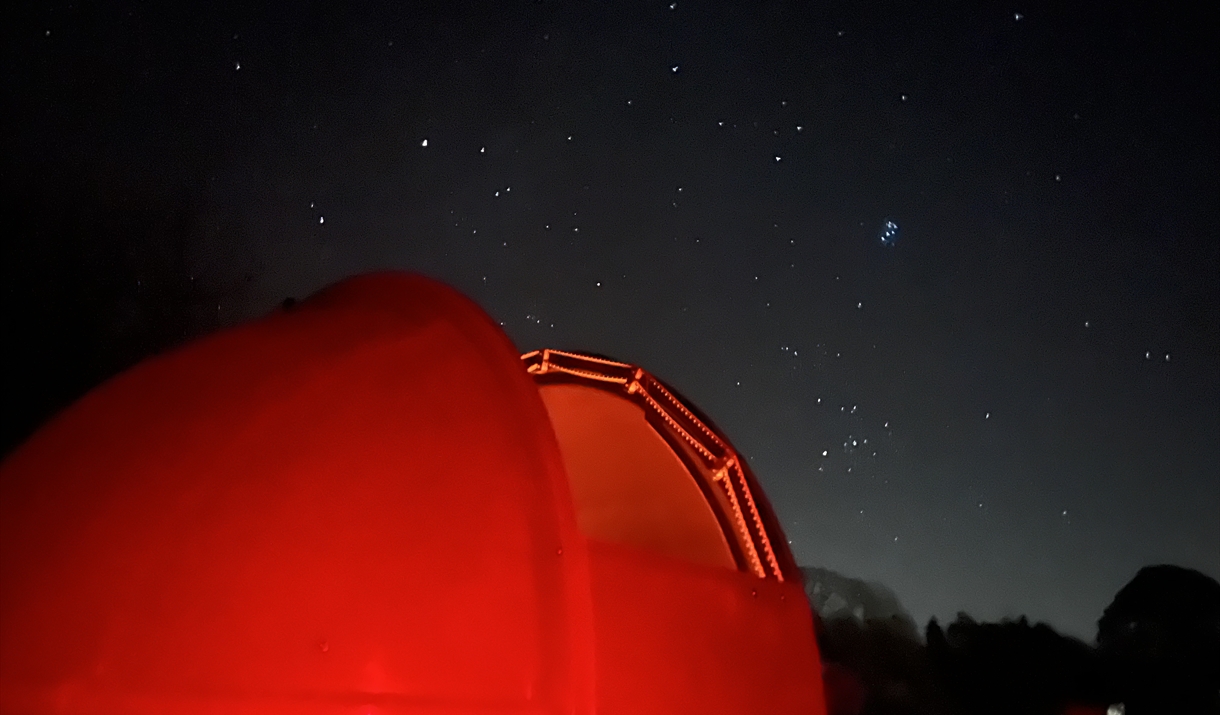 A red-lit observatory dome at night beneath a star-filled sky, with a dark landscape and trees visible in the background.