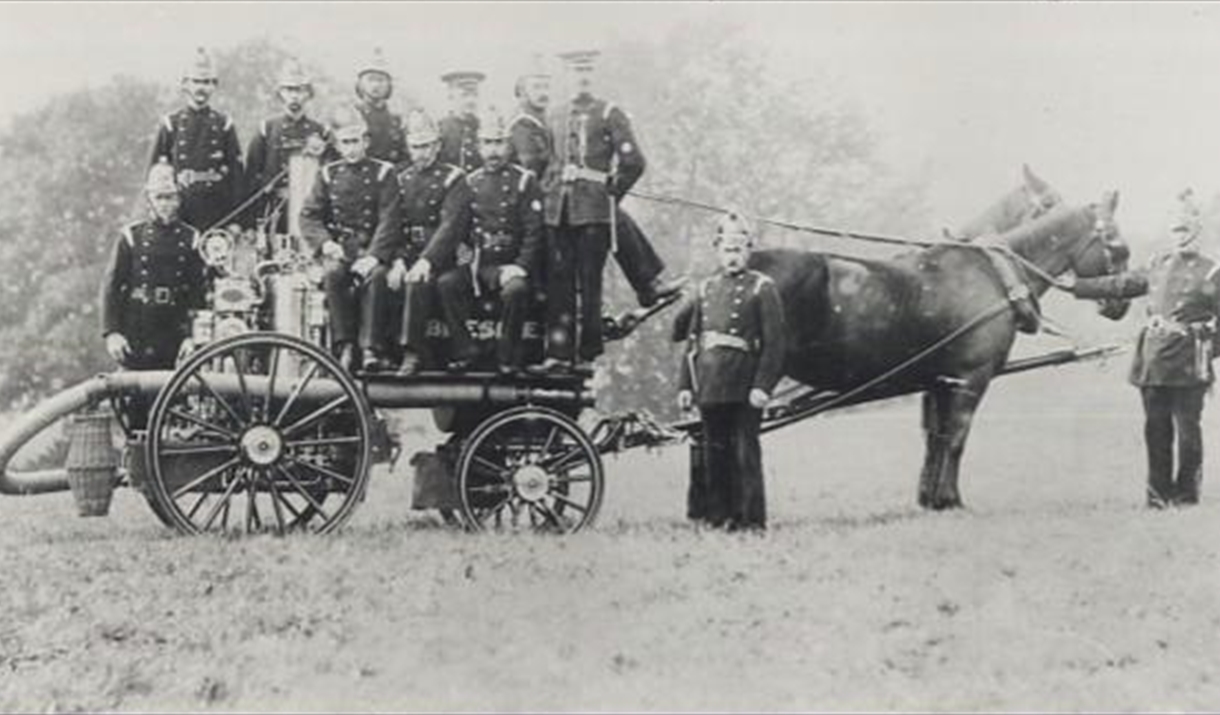 Image of fire fighters sat on a trailer with a horse connected to the front. The trailer contains a hose and a water pump system.