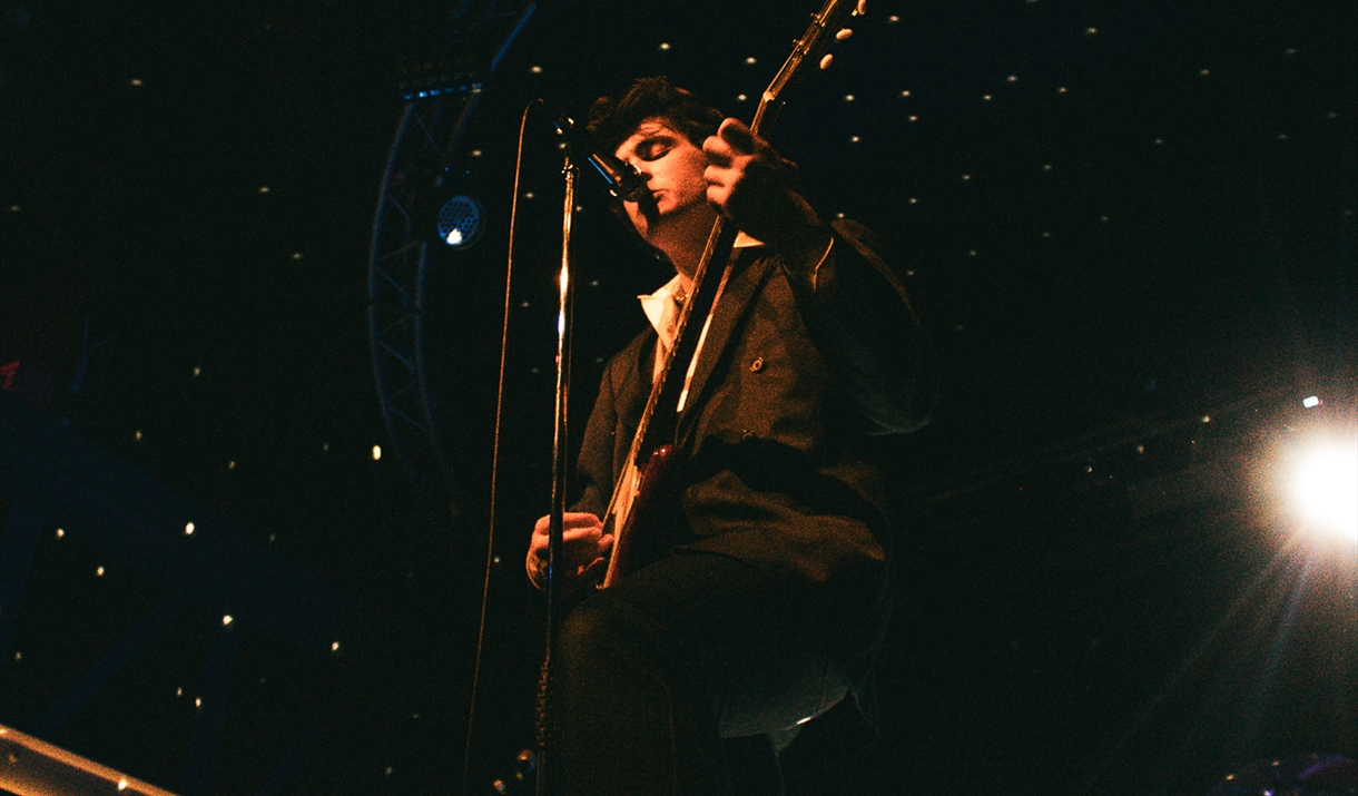 Lead singer of arctic numpties plays guitar on stage with lights behind him