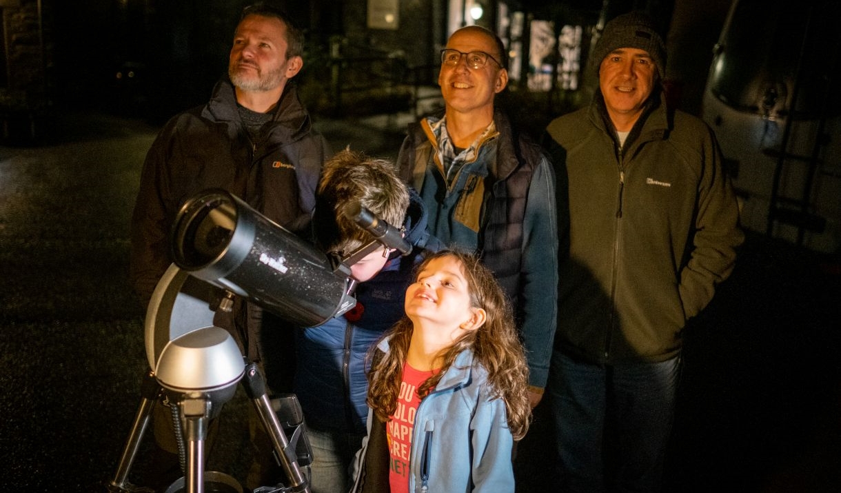 Two children looking through a telescope at night accompanied by 4 adults
