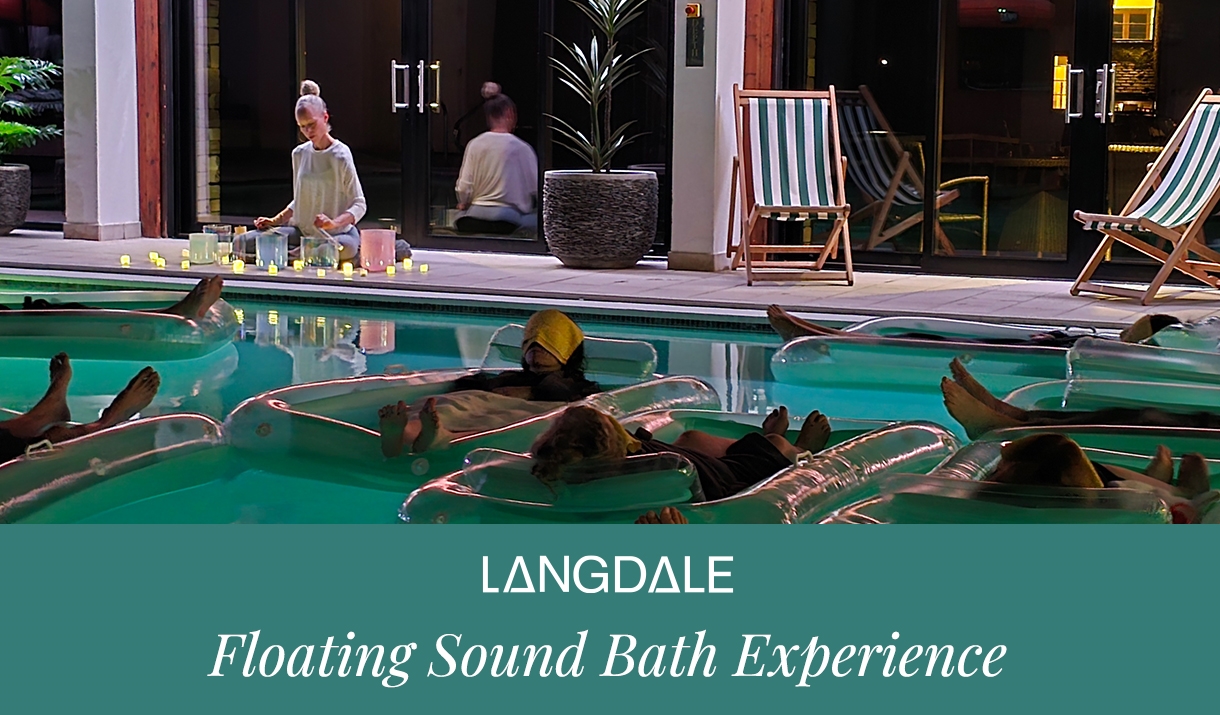 People relaxing on floating mats in a pool during a nighttime sound bath session, while an instructor plays crystal singing bowls beside the water at
